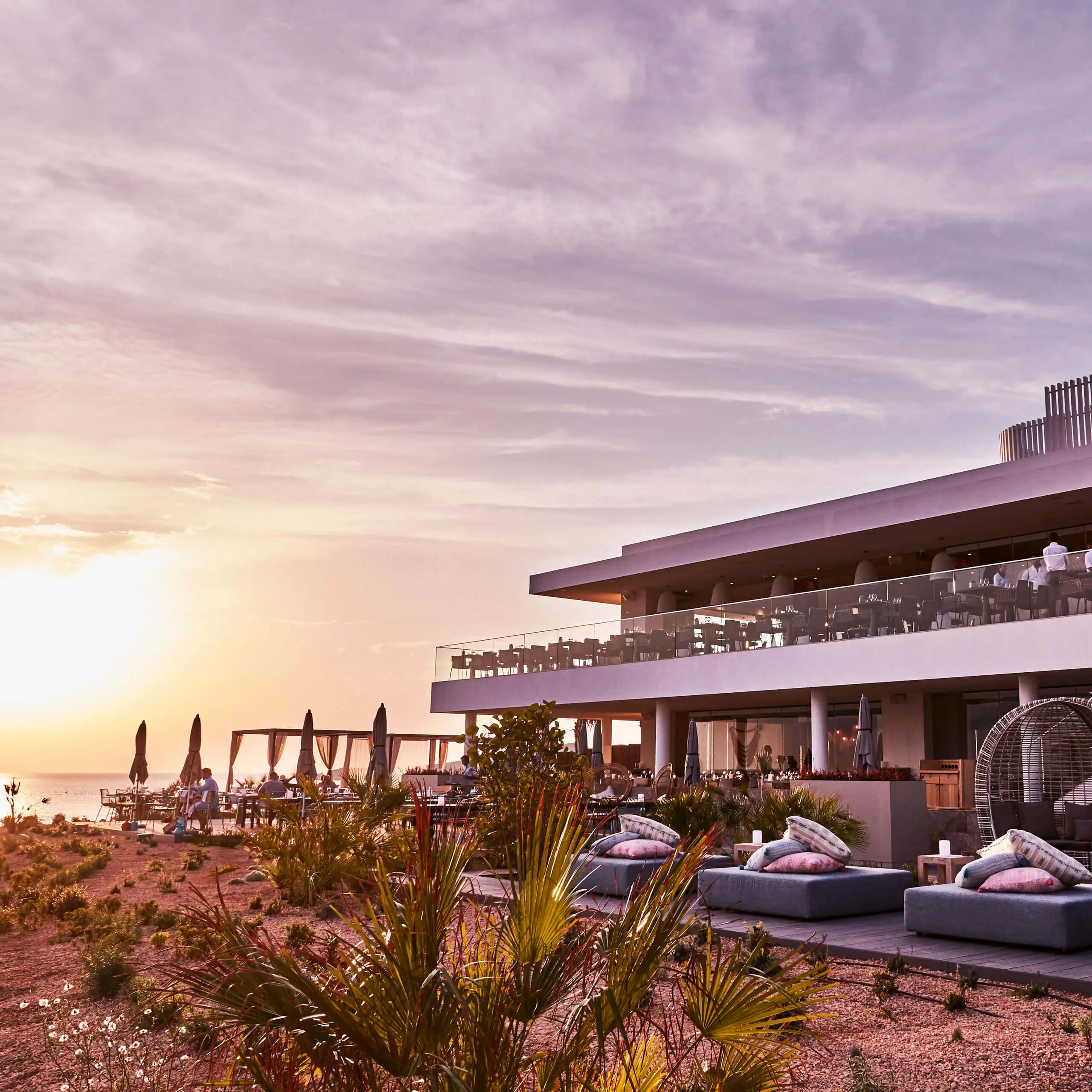 Terrasse des Nela Pop-up Restaurants im 7Pines Hotels & Resorts mit Tischen im Freien, Lounge-Bereich und Meerblick bei Sonnenuntergang.