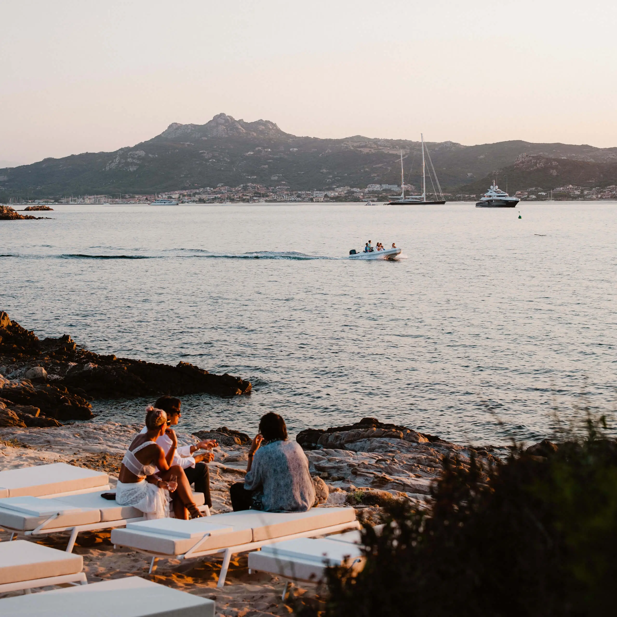 People relaxing on a rocky beach with boats at sunset.