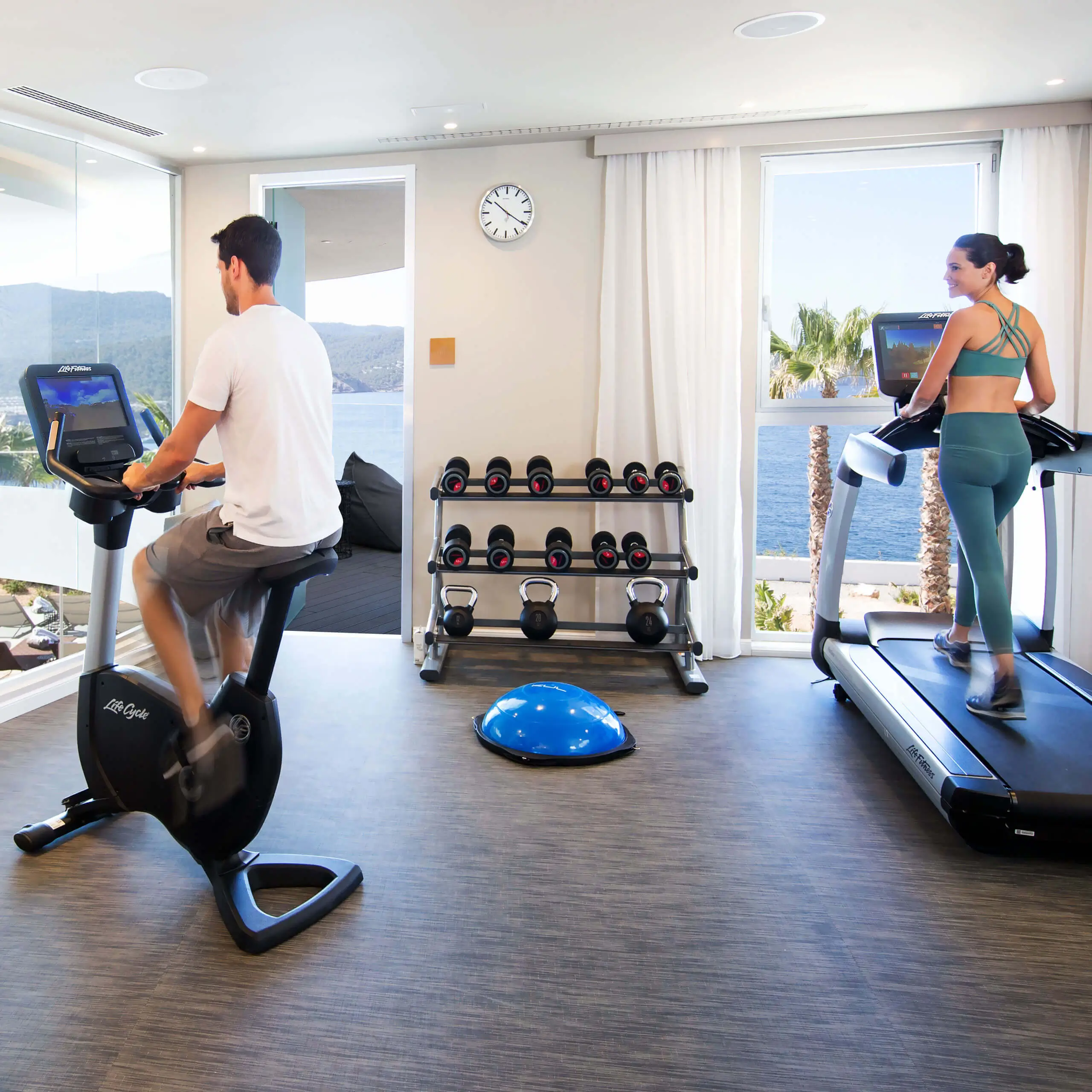 Heller moderner Fitnessraum in den 7Pines mit Heimtrainer, Laufband, Hanteln und Meerblick.