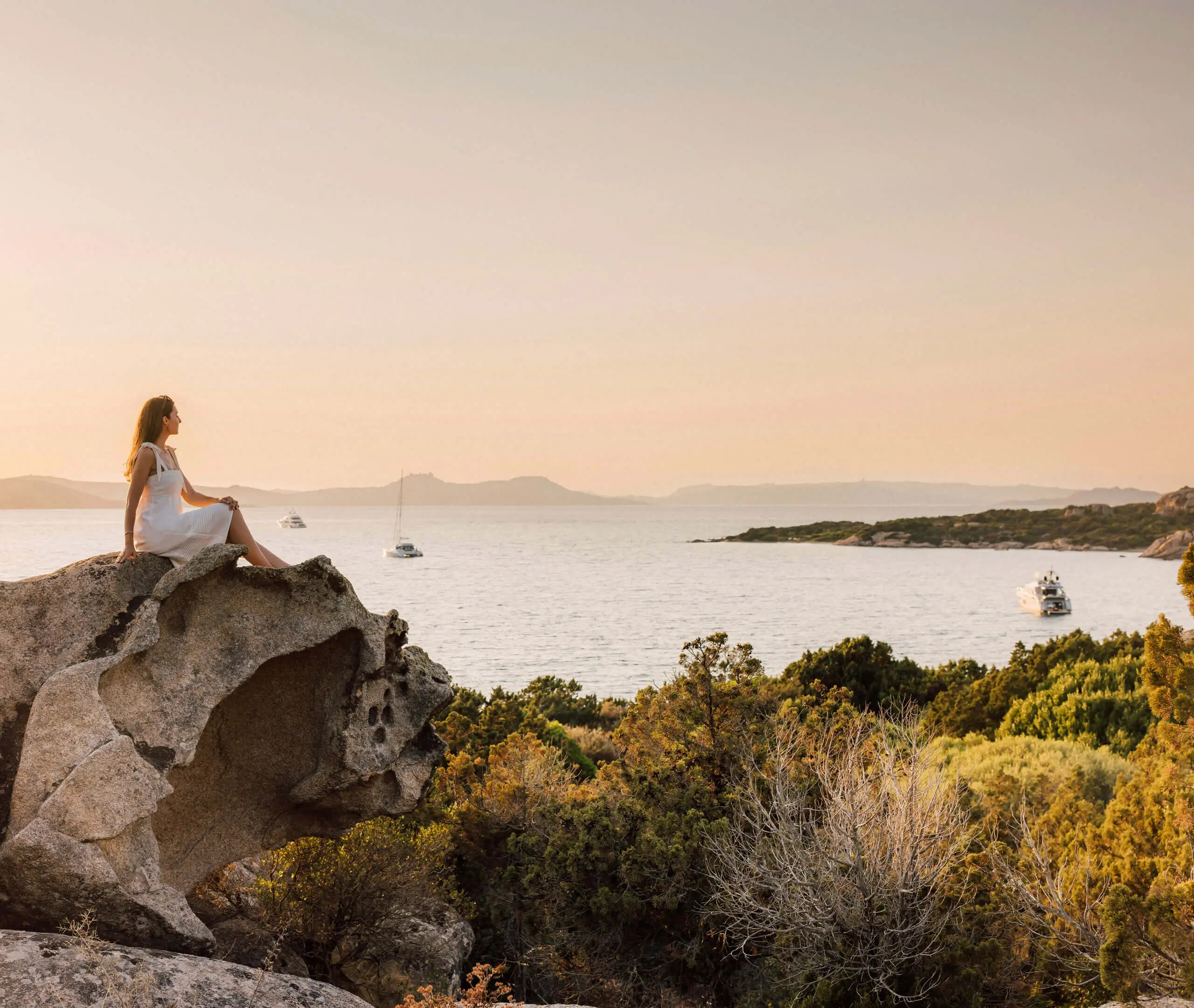 Frau im weißen Kleid sitzt bei Sonnenuntergang auf einem Felsen und blickt bei 7Pines auf das Meer und Boote.
