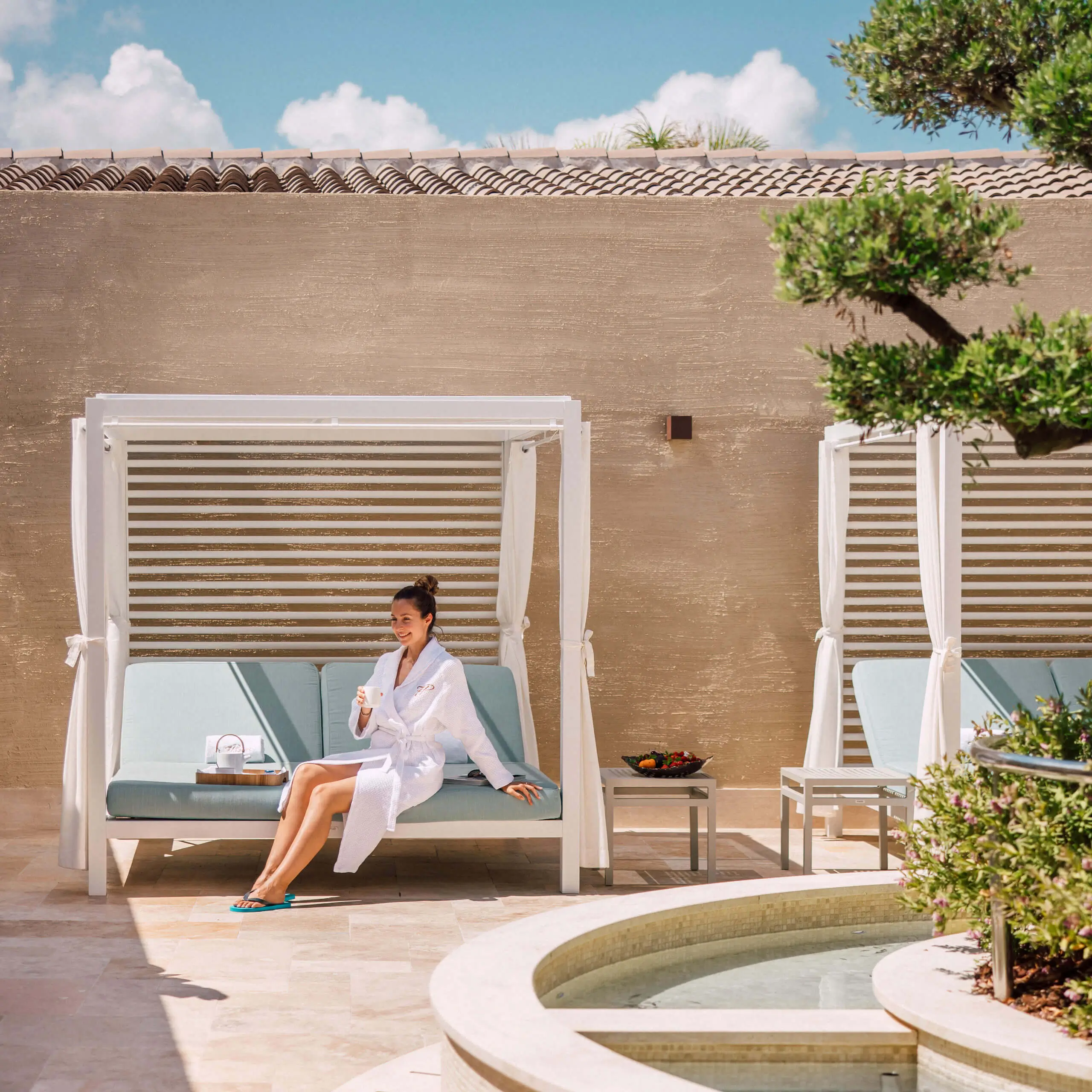 Eine Frau entspannt sich in einer Cabana am Pool eines luxuriösen 7Pines Resorts und hält ein Getränk neben frischem Obst.