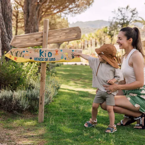 Mother and child at 7Pines Resorts pointing to KIO - House of Kids sign in a green outdoor setting.