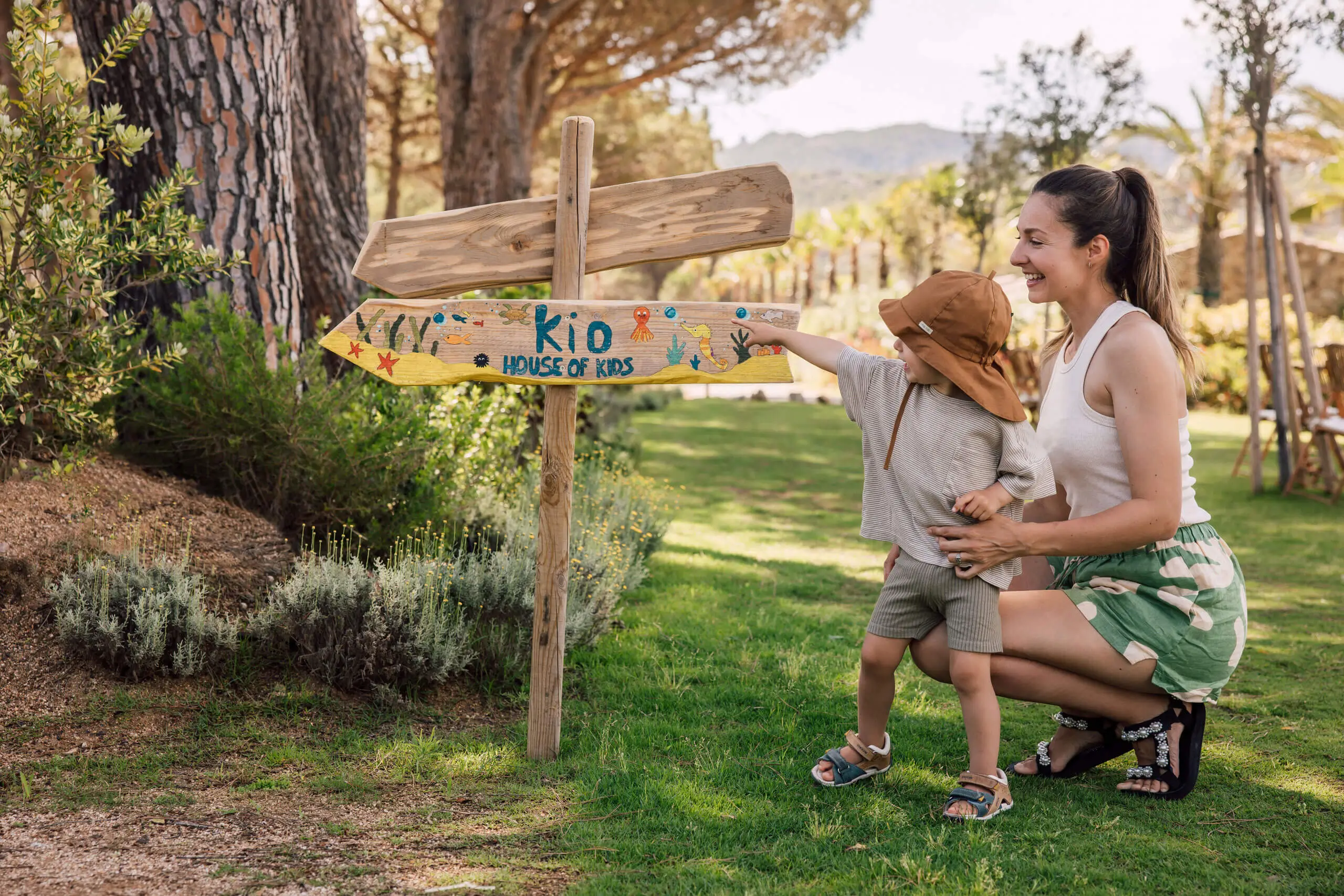 Eine Mutter und ein Kind am Kio House of Kids-Schild in einem üppig grünen Bereich.