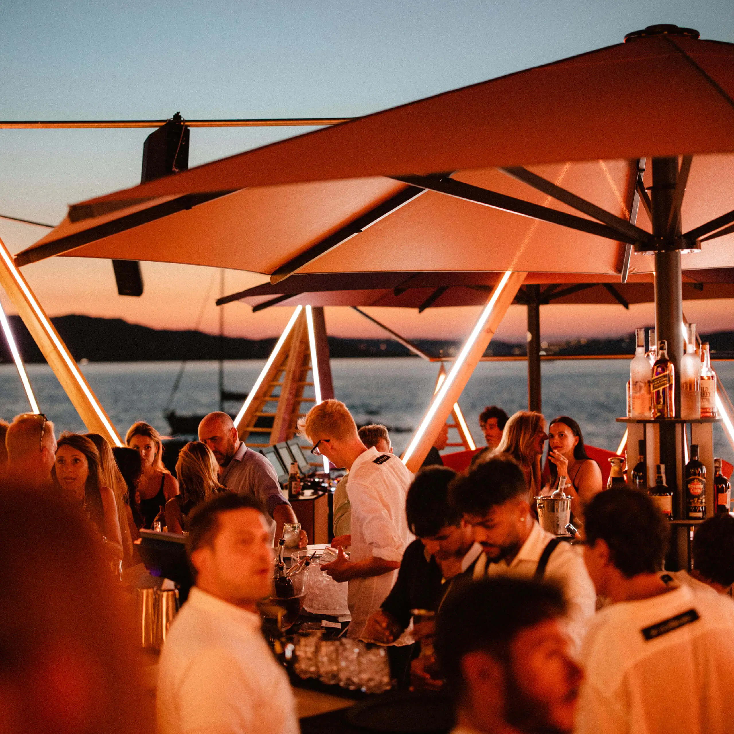 Guests and bartenders at Cone Club beach bar at sunset, with illuminated umbrellas, cocktails and the sea in the background.