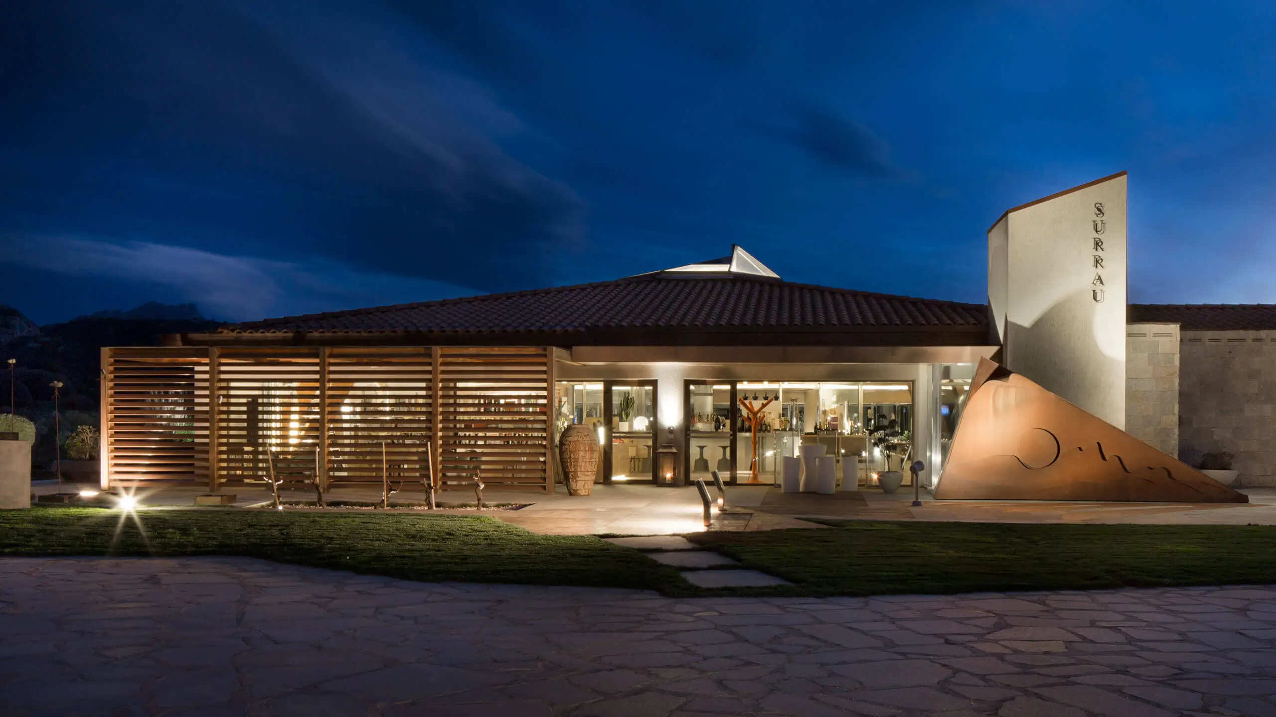 Night view of Surrau restaurant at 7Pines Hotels & Resorts, with an illuminated modern facade and lawn.