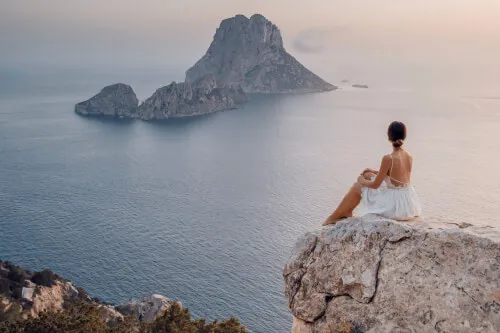 A woman in a white dress sitting on a rock by the sea with an island in the background.